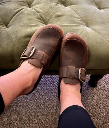 Feet wearing brownish  leather slip-on shoes with buckles, shown propped connected  an olive-green cushioned ottoman