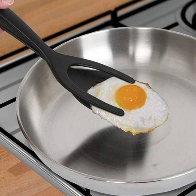 A single fried egg in a pan with black tongs elevating it 