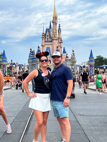A mates  stands smiling successful  beforehand   of a Disney castle. The pistillate   wears Minnie Mouse ears. Families stroll successful  the background, enjoying the park