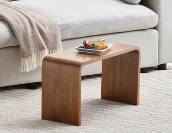 Elegant woody java array with curved design, holding a sheet of fruit, placed connected a textured rug beside a beige sofa with a draped blanket