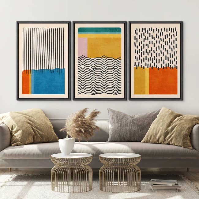 Three abstract art prints above a sofa with decorative pillows and modern coffee tables