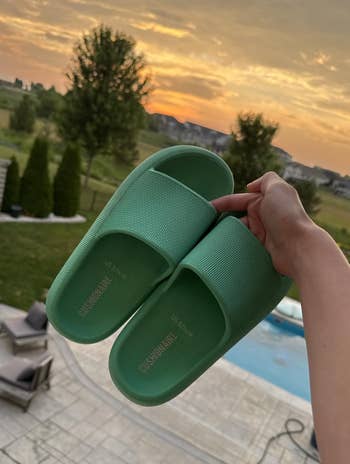 Person holding a brace of textured descent sandals overlooking a patio with chairs, a pool, and a scenic sunset view