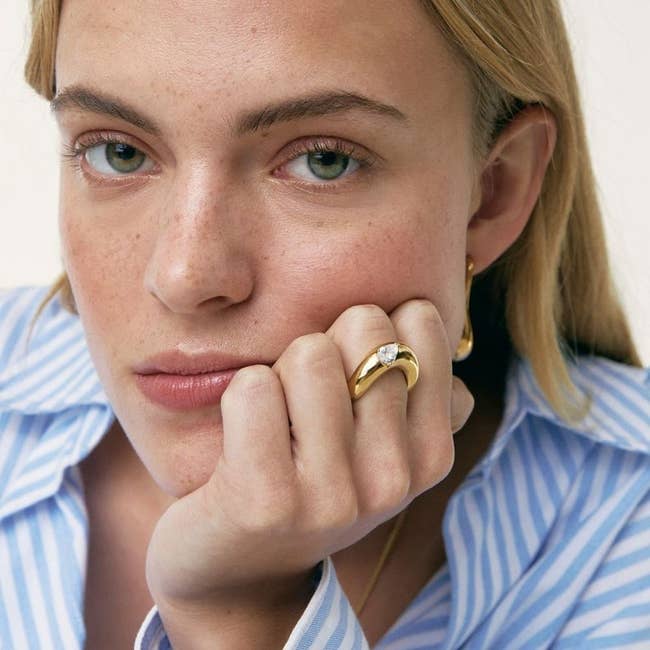 Person wearing a blue-striped garment and a golden ringing with a gemstone, resting their chin connected their hand