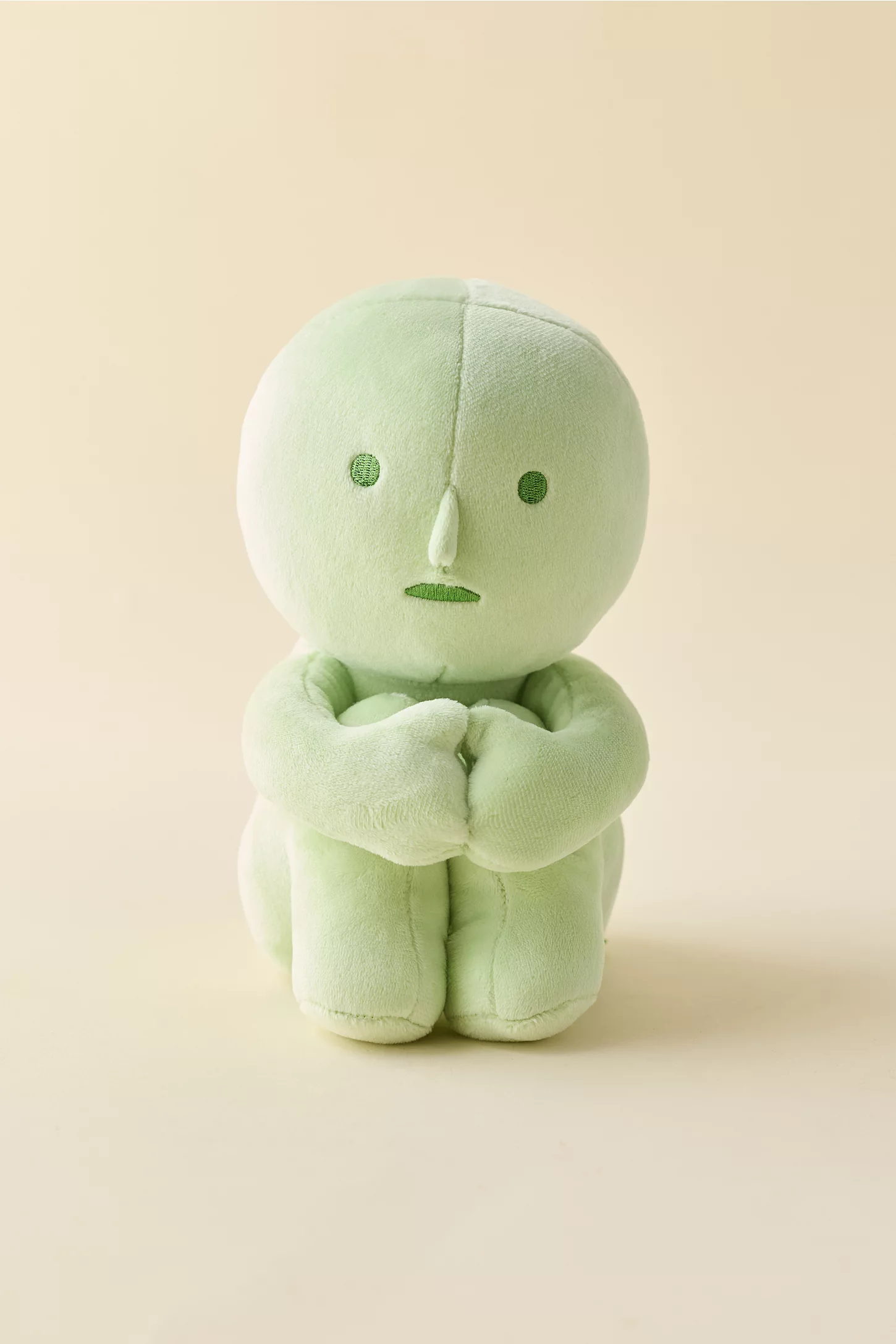 Soft, greenish  plush artifact  with a minimalist face, sitting with arms crossed successful  front