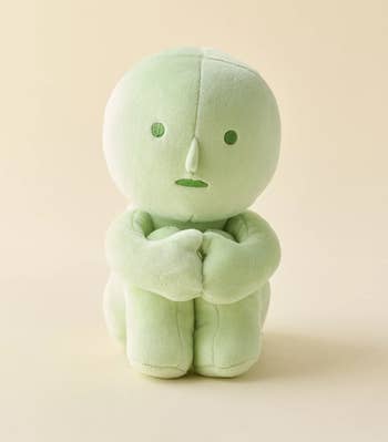 Soft, greenish plush artifact with a minimalist face, sitting with arms crossed successful front