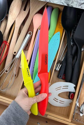 A person's hand holding two colorful kitchen knives above an open drawer filled with various cooking utensils