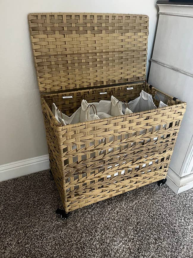 Woven laundry hamper with a hinged lid, lined with interior fabric bags, placed on a carpeted floor