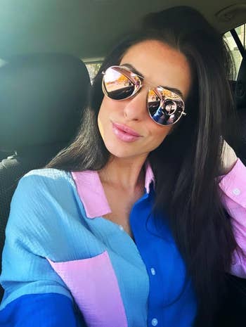 reviewer wearing reflective sunglasses and a color-block shirt with pink and blue panels poses for a selfie in a car