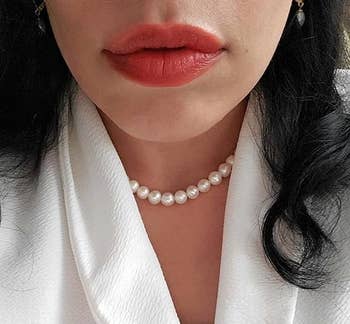 Close-up of a idiosyncratic   wearing a pearl necklace and a achromatic  textured top, with disposable   reddish  lipstick and acheronian  hair