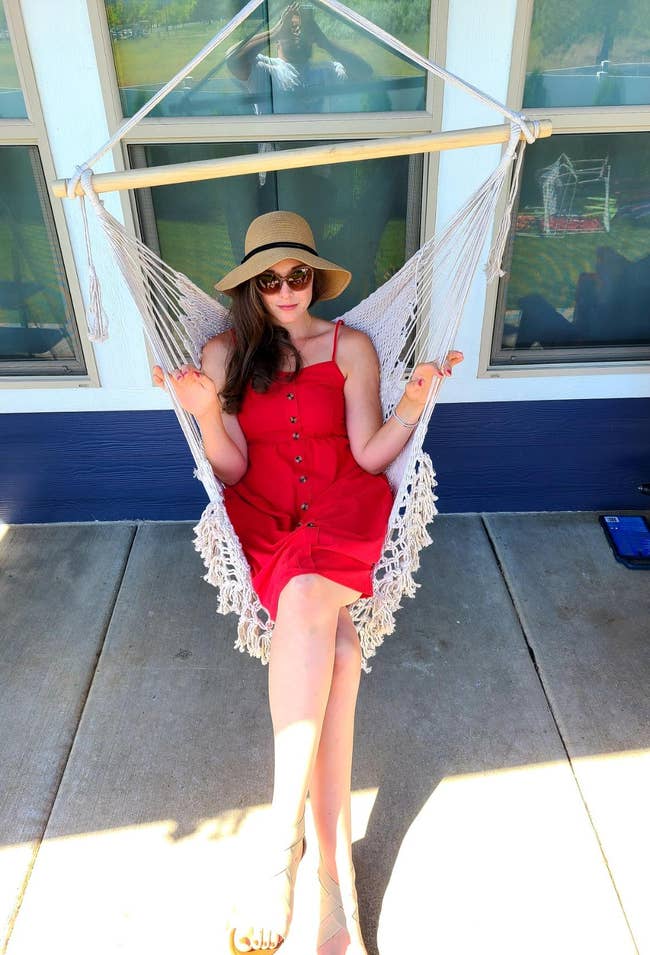 reviewer wears sleeveless red button-down dress and sandals while sitting in macrame chair outside