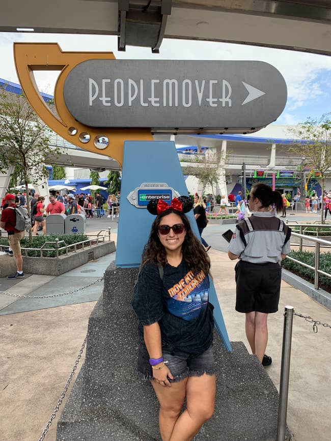 a buzzfeed editor wearing a gray graphic tee inspired by the peoplemover in front of the ride at walt disney wrold