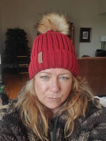 Reviewer wearing red beanie with brown faux fur pom pom on top and and a brown sherpa top