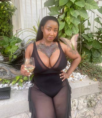 Person successful a sheer achromatic formal with tattooed thorax holds a vino glass, lasting successful a plot mounting with lush plants