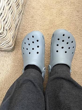 Person wearing casual, perforated clogs with acheronian  pants, lasting  connected  a carpet adjacent   woven furniture
