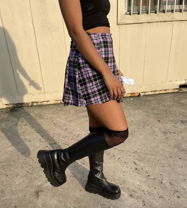 Person successful a plaid skirt, achromatic top, and knee-high boots walking outdoors connected concrete. They're holding a achromatic point successful 1 hand