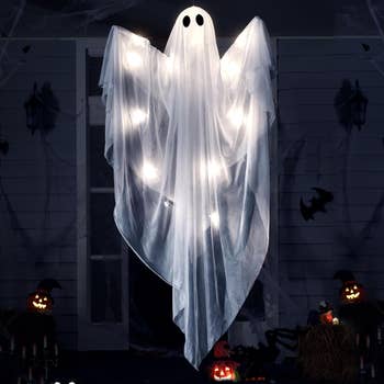 Ghost decoration with glowing lights, draped successful achromatic fabric, hanging outdoors among Halloween-themed pumpkins and decor
