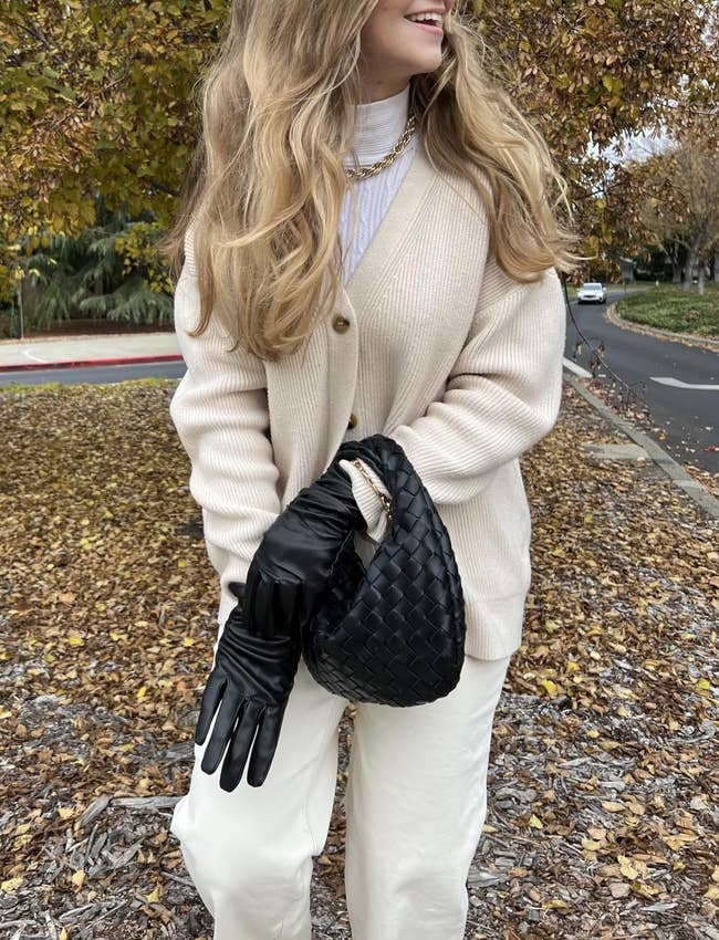 Reviewer successful a cozy cardigan with a stylish woven achromatic handbag and matching gloves, posing outdoors among autumn leaves