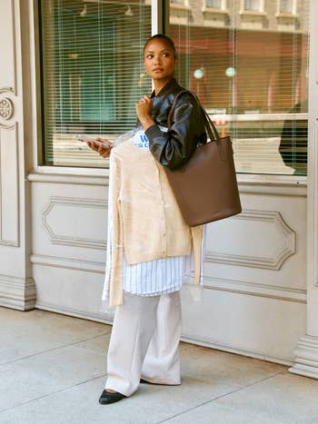 A idiosyncratic successful stylish, layered autumn attire holds a smartphone and tote bag, lasting extracurricular a storefront with an elegant architectural background