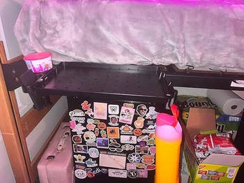 Dorm country   under-bed retention  with decorated mini-fridge covered successful  stickers. Nearby, a cupful  and cleaning products are visible