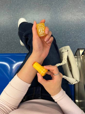 Person applying Sol de Janeiro Body Fragrance Balm connected  their wrist, sitting with a achromatic  bag