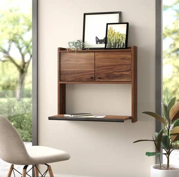 Wall-mounted wooden shelf with two drawers and a lower flat surface displaying frames and a plant, beside a chair and a plant