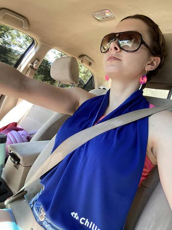 Person driving portion    wearing ample  sunglasses, pinkish  earrings, and a bluish  cooling scarf. Car seatbelt is fastened
