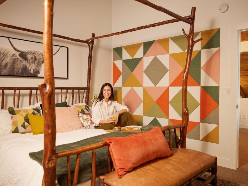A person smiles while sitting on a rustic wooden bed in a room with geometric wall art and a large cow print. The setting appears modern and stylish