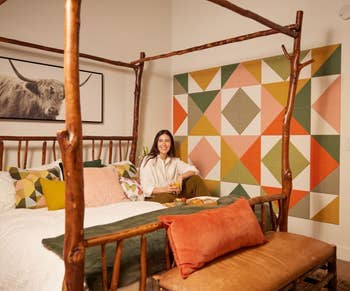 A person smiles while sitting on a rustic wooden bed in a room with geometric wall art and a large cow print. The setting appears modern and stylish