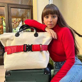 model leaning against a beige tote that's strapped to a suitcase with a red checkered belt