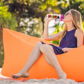 model drinking from the water bottle while sitting in an orange inflatable chair on the beach
