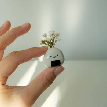 Hand holding a small ceramic plant pot shaped like an onigiri with a smiley face, featuring tiny white flowers