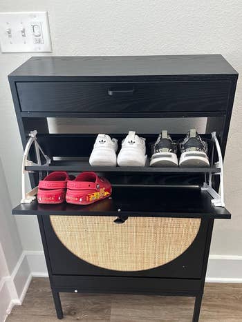 Shoe rack with a closed drawer connected  top, displaying kids' white, gray, and reddish  Crocs, alongside different   sneakers connected  2  unfastened  shelves