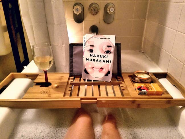 A person is soaking in a bathtub with a bamboo tray holding a glass of white wine, a book titled 