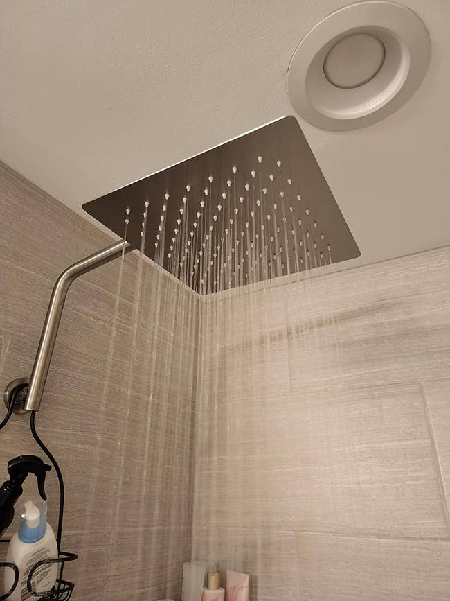 Large square showerhead with water streaming down in a modern bathroom setting. Various bath products are seen below the showerhead