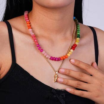 A idiosyncratic   wearing a achromatic  apical  showcases a colorful beaded necklace paired with a golden  pendant necklace