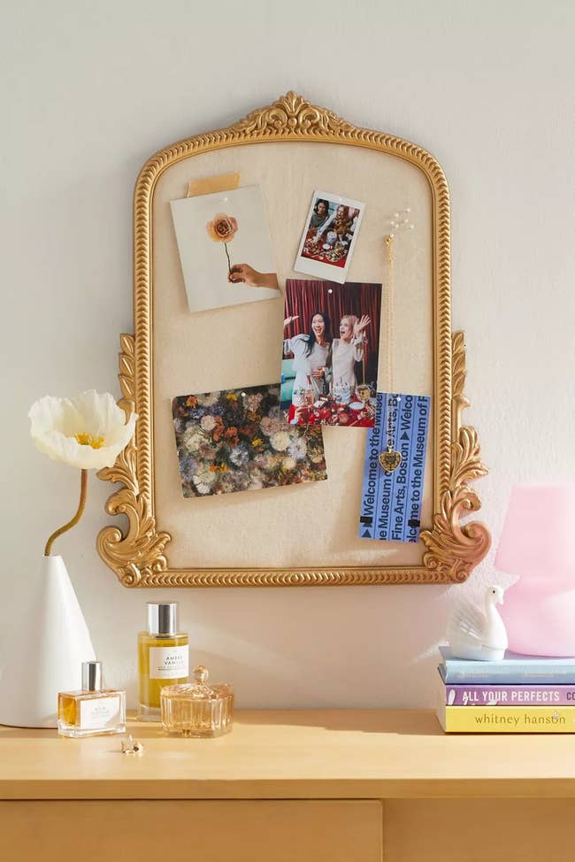 Ornate gold frame corkboard with photos, perfumes, a flower, and a ribbon on a dresser