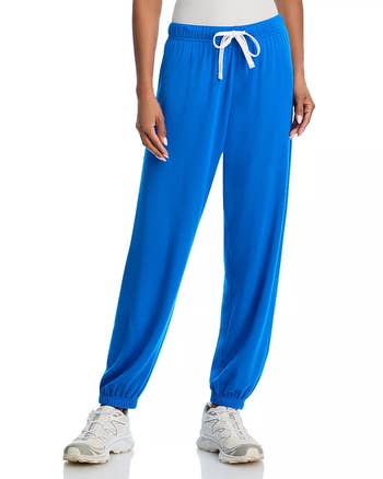 Person wearing bluish  joggers and achromatic  sneakers stands against a plain background