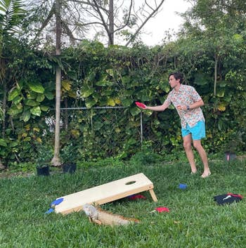 Person playing cornhole game in a backyard, preparing to toss a beanbag. Wearing a patterned shirt and shorts