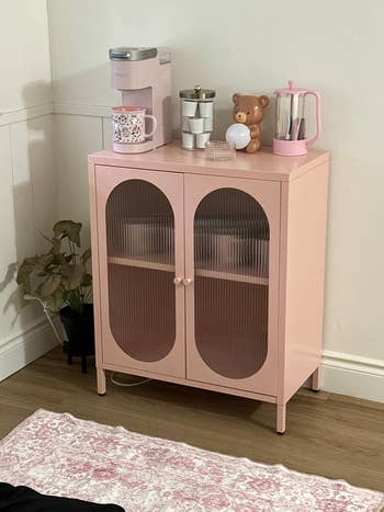A pinkish  furniture  with ribbed doors, topped with a java  maker, mug, and decor; beside it is simply a plant. Cozy room  oregon  eating  country  setup