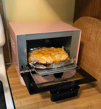 A small, pink countertop toaster oven baking a dish with melted cheese on top of a bread