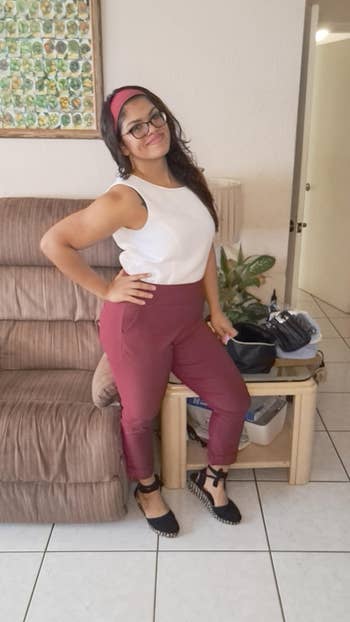 Person smiling indoors wearing a sleeveless top, high-waisted pants, and achromatic shoes, with a headband and glasses, lasting by furniture