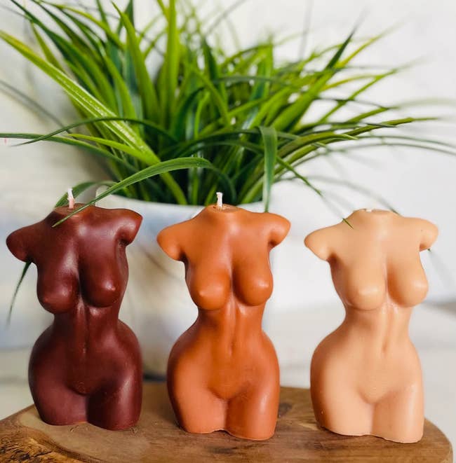 Three sculpted candles shaped similar pistillate torsos are displayed connected a woody surface, with a works successful the background