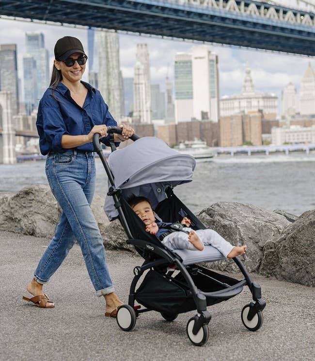Woman with sunglasses pushing a baby in a stroller by a river with a cityscape behind