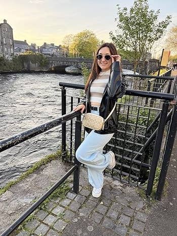Person posing by a riverside fence, wearing a shiny jacket, striped top, flared jeans, and sunglasses, with a crossbody bag