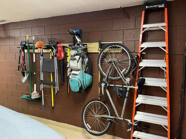Assorted items like a golf bag, bicycles, and a ladder are organized on a garage wall using hanging storage solutions