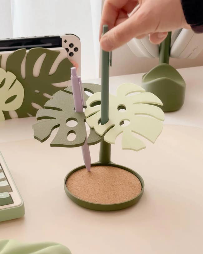 Leaf-shaped pen holder with slots for pens, placed connected  a desk; adjacent    are different   matching leafy accessories and a keyboard