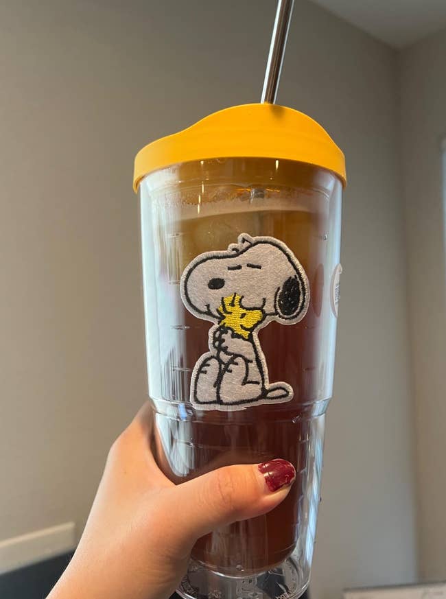 A manus holding a ample tumbler with a Snoopy and Woodstock design, filled with iced beverage and a metallic straw