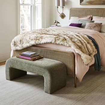 A chic bedroom showcasing a neatly made bed with a faux fur throw, layered pillows, and a green velvet bench at the foot. Elegant wall sconces and books enhance decor