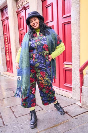 Person in eclectic outfit with patterned jumpsuit, green scarf, black hat, and boots, posing confidently on a street