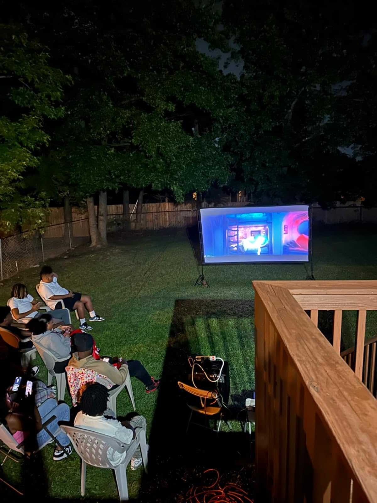 Outdoor movie   nighttime  setup with respective  radical   seated successful  chairs watching a projected movie  successful  a backyard. Trees and a obstruction   are visible